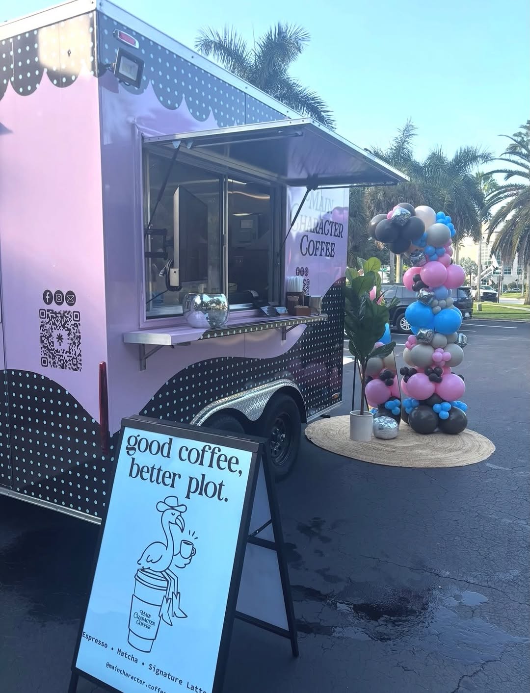 The Main Character Coffee trailer set up and serving at its grand opening: serving window open, pink and blue balloons, and the A-frame sign reading good coffee, better plot